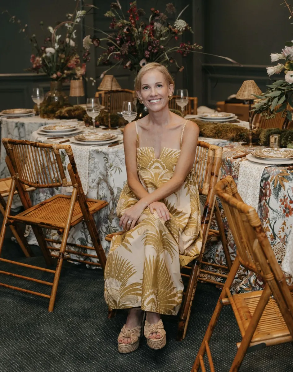 Ashley Snider smiling, seated in a golden palm print dress among bamboo chairs and a dressed tablescape