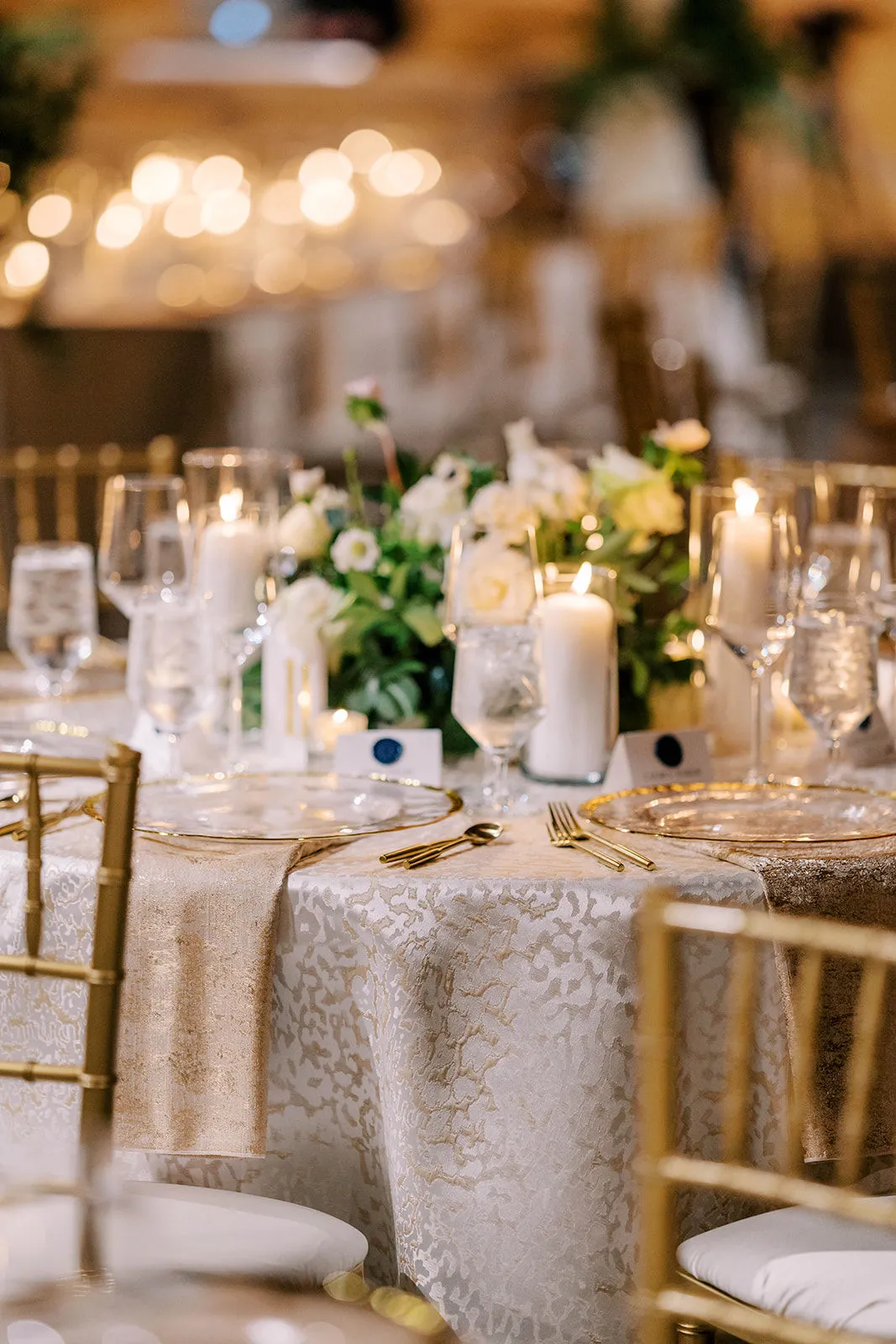 A candlelit reception table of white roses and anemones on gold jacquard linen with gold rimmed chargers and gold flatware